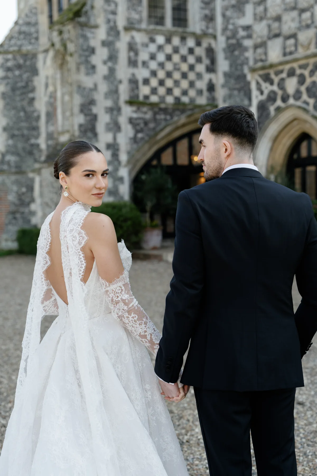 Woman in lace bridal gown looks over her shoulder while walking with a man in a tuxedo. Soft, neutral background. She has a calm, poised expression.