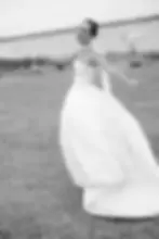 Bride in a flowing gown twirls joyfully on a grassy field by a lake. She holds a glass, smiling, with boats in the distant water. Black and white.
