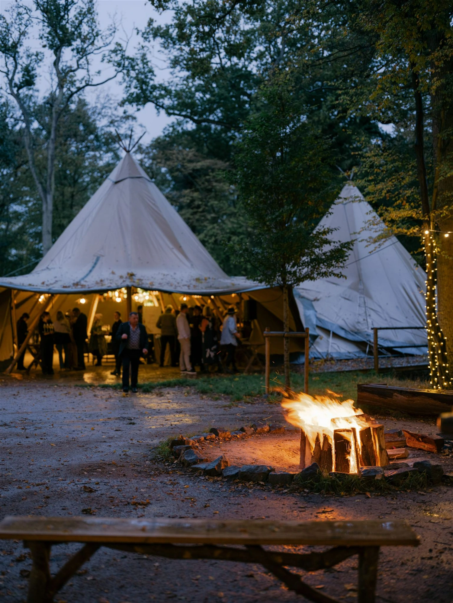 A view of giant hat tipis against a dusky sky with a bonfire in the foreground.