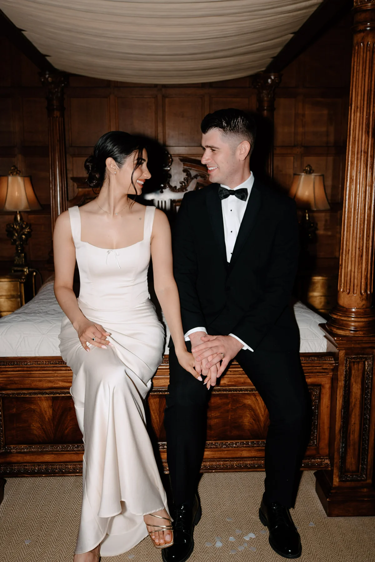A bride and groom sit on the edge of their hotel bed before their reception, dressed in formal attire, holding hands and smiling.