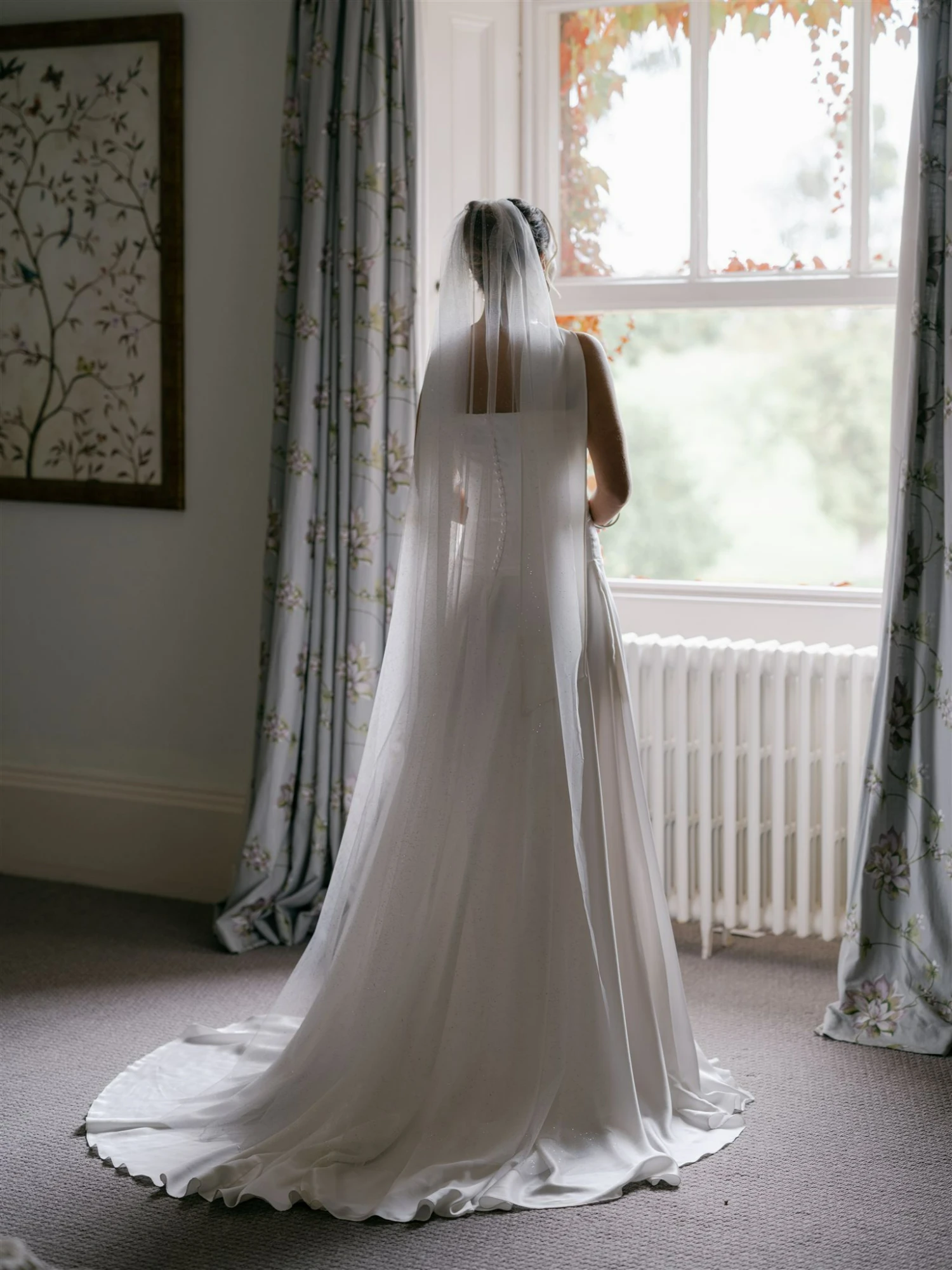 A bride facing a window wearing a long wedding dress and veil - there is a soft glow to the image.
