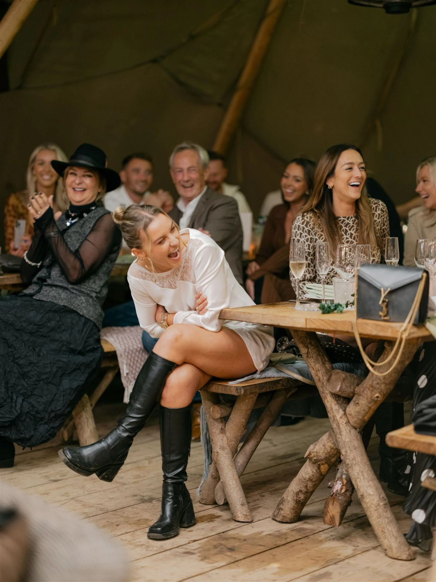 A woman wearing black boots and a short white dress is seated at a table and laughing while guests around her smile and laugh.