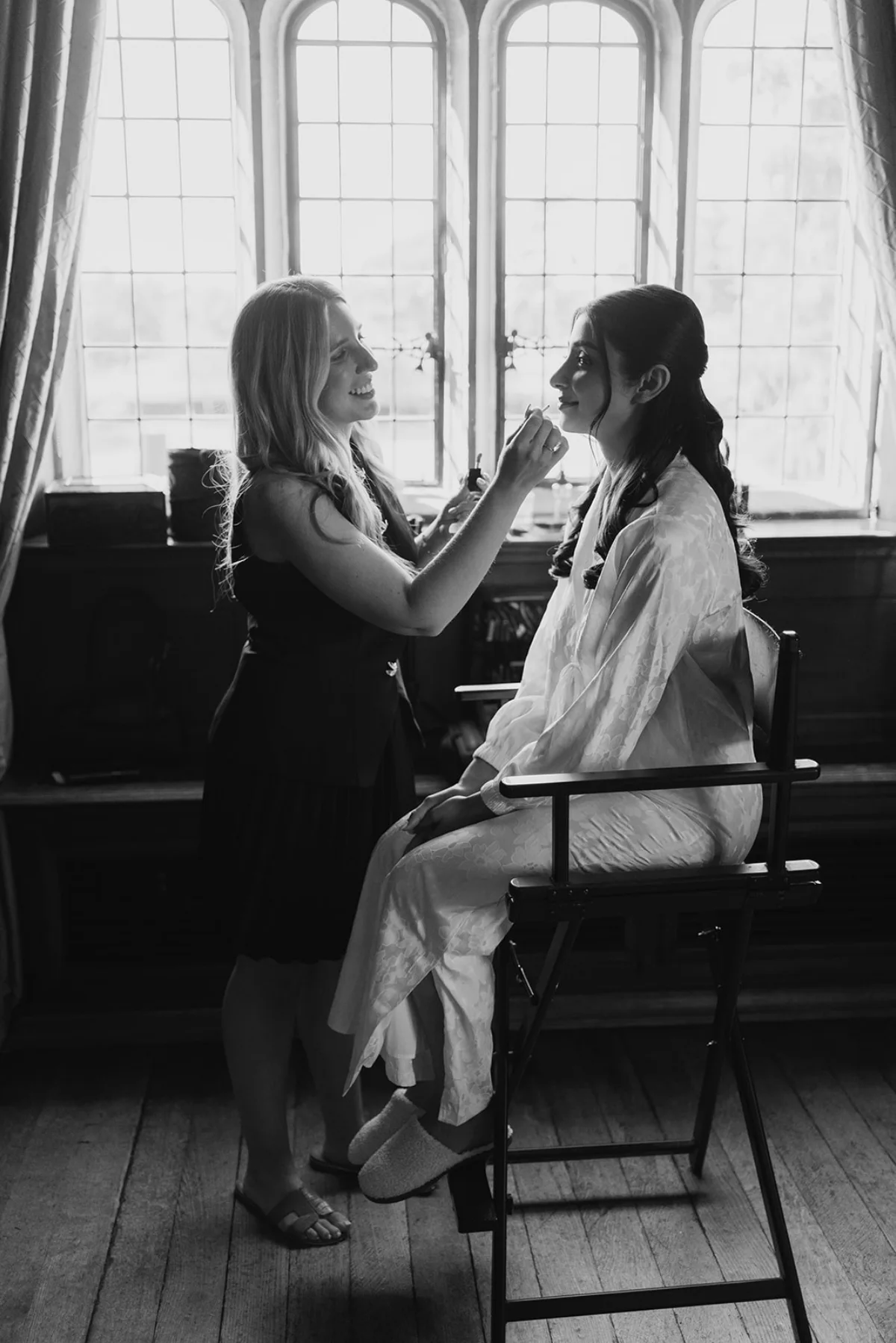 Suffolk bridal makeup artist Emily Chantal preps a bride on her wedding day against a window with bright light. Black and white photo.