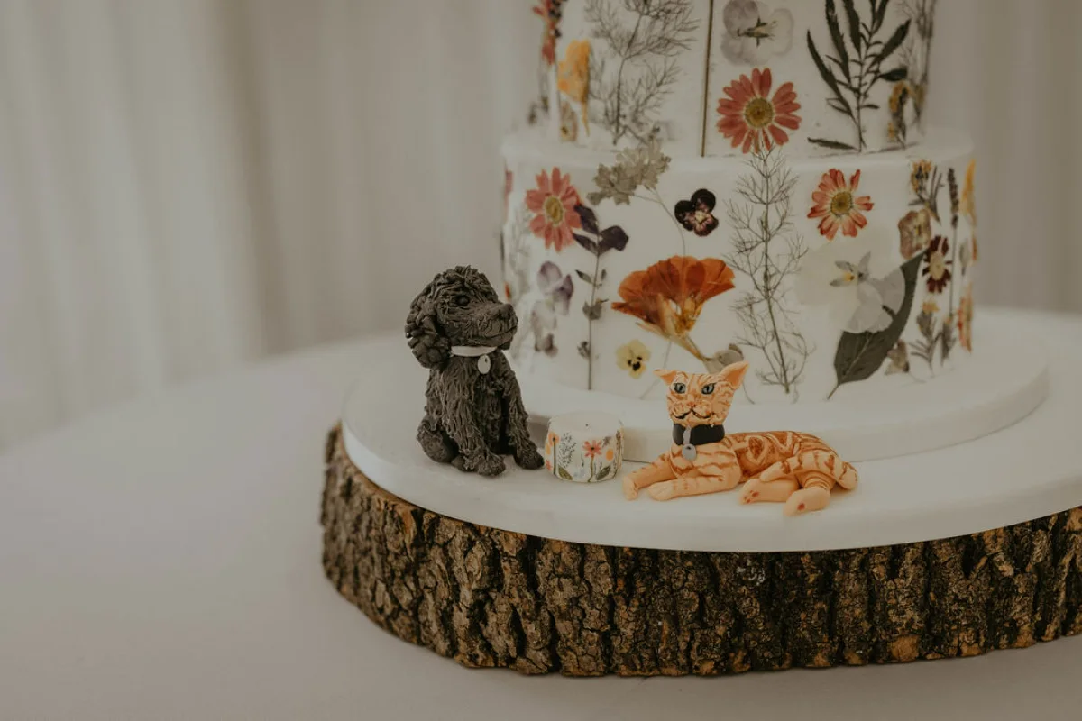 A photo of a wedding cake featuring dried flowers pressed on the sides, and a fondant black spaniel dog and ginger cat next to a smaller fondant version of the cake. Suffolk wedding photography by Mikaela Jade.