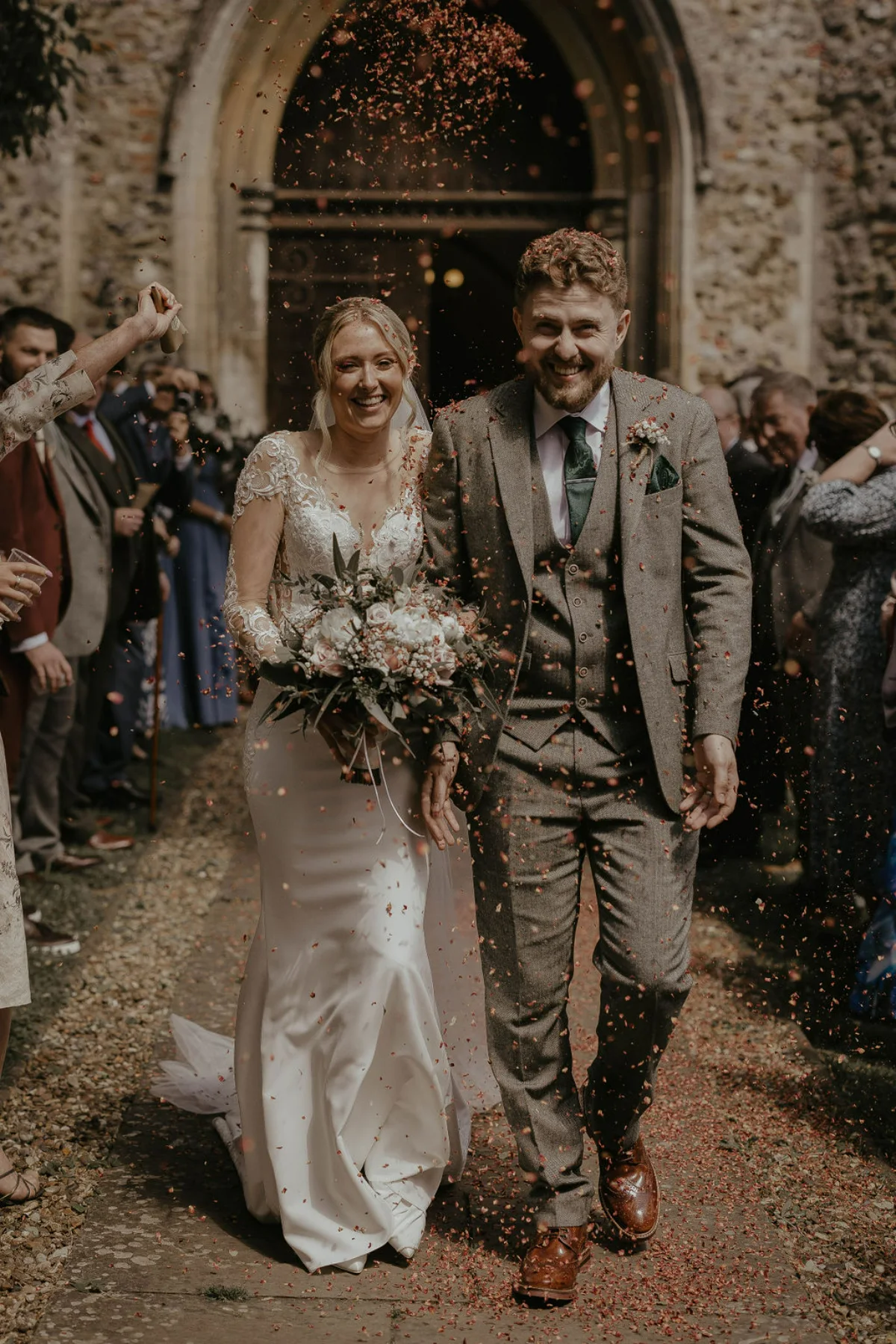 A bride and groom exit a church to wedding guests throwing confetti. Happy and joyful image. Suffolk wedding photography by Mikaela Jade.