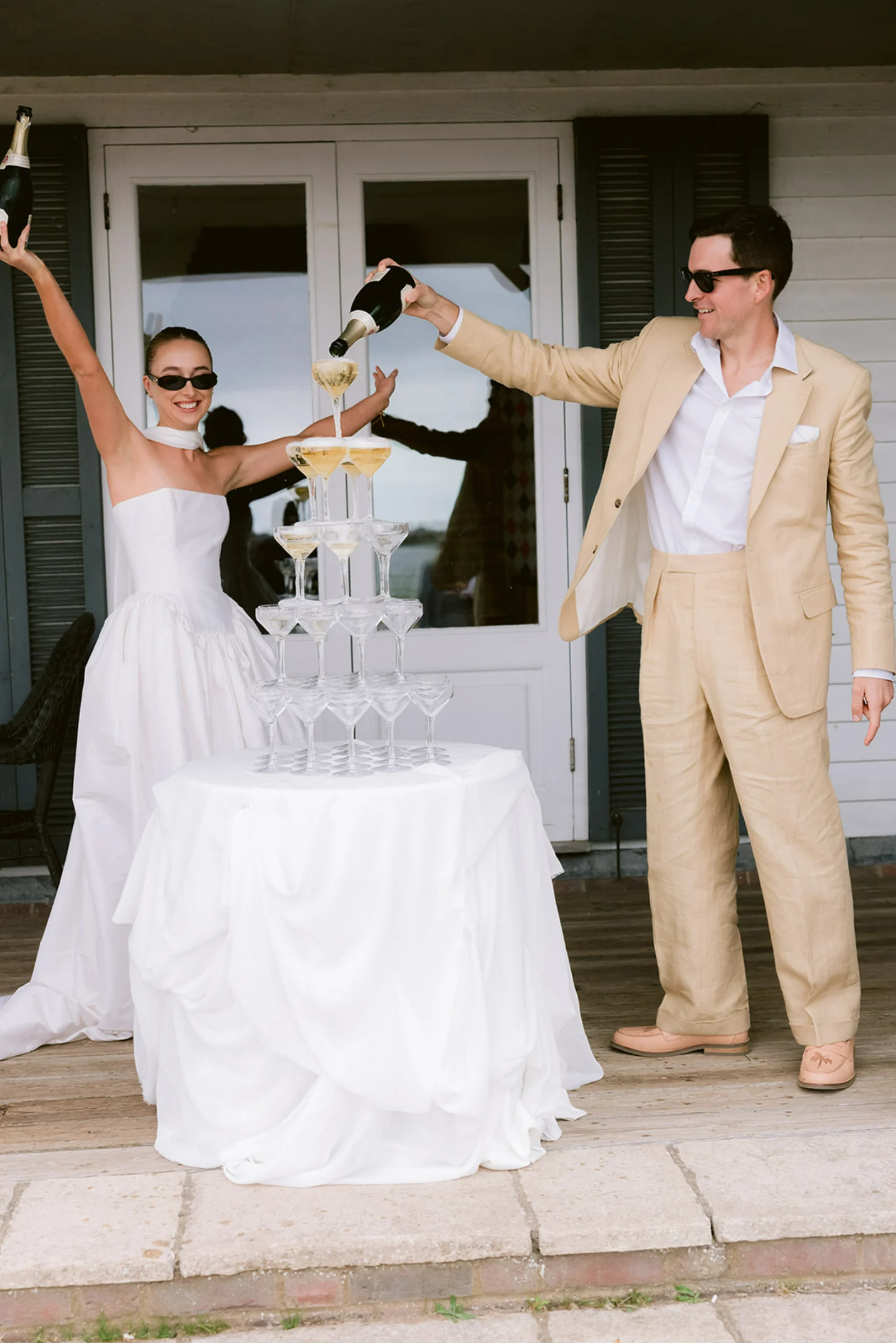 A bride and groom pour Champagne into a tower of glasses. The bride throws her hands into the air in celebration.
