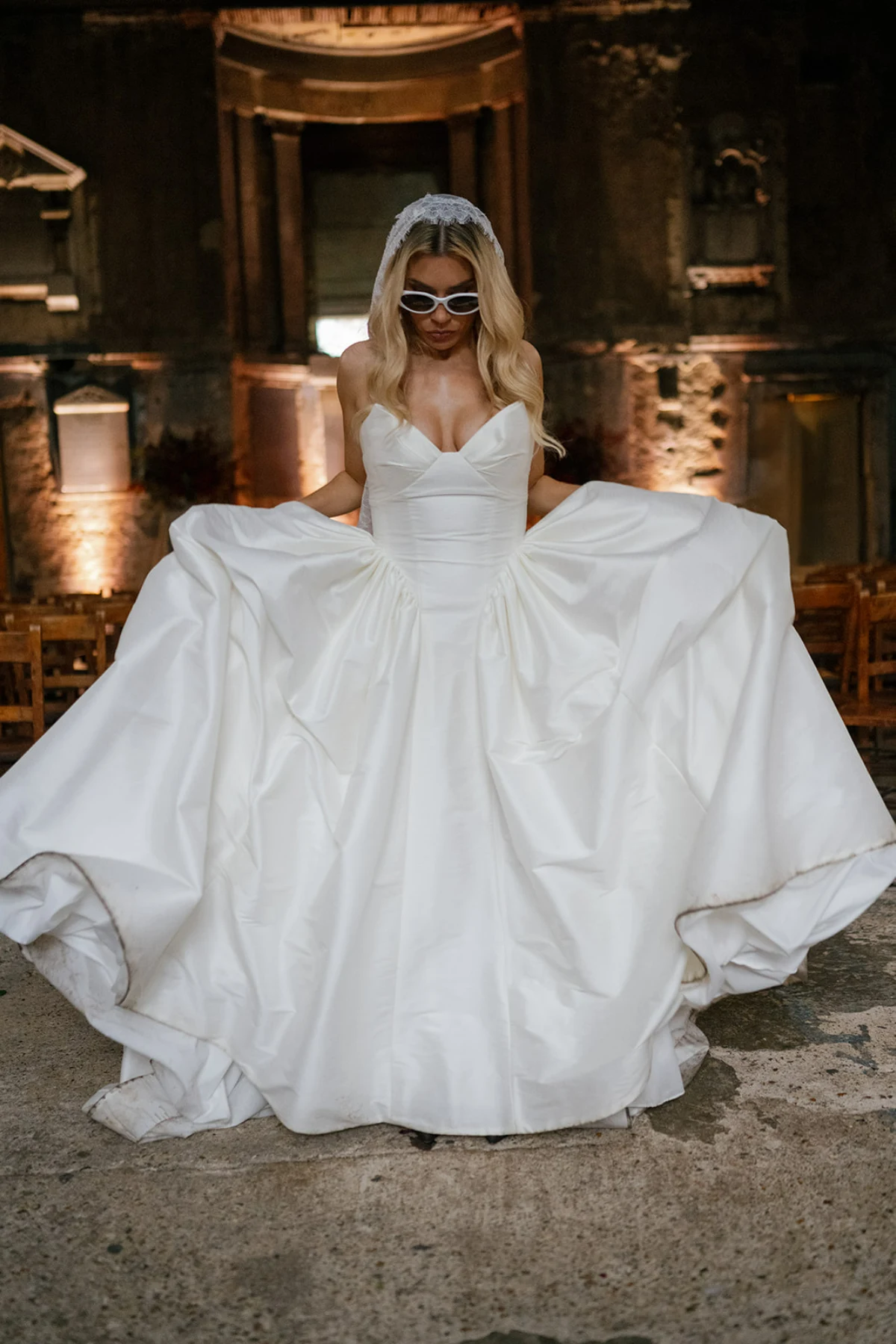 A bride wears sunglasses, posing by fluffing up her dress inside Asylum Chapel.
