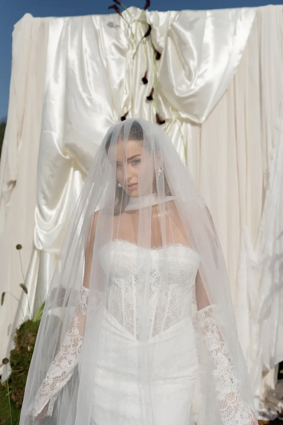 A bride in a lace gown and veil stands before white draped fabric with dark floral accents, set against a clear blue sky.