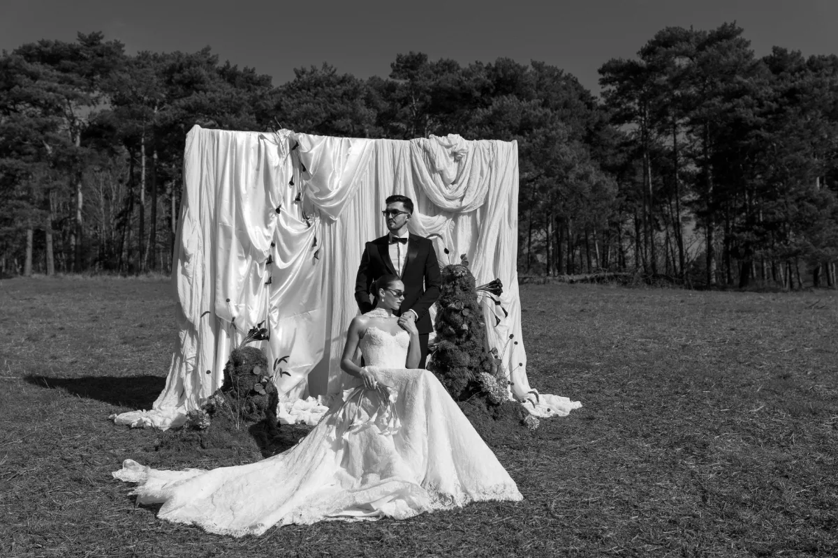 A bride and groom wear sunglasses and pose together in front of a backdrop of white fabrics, wearing makeup by Suffolk makeup artist Emily Chantal.