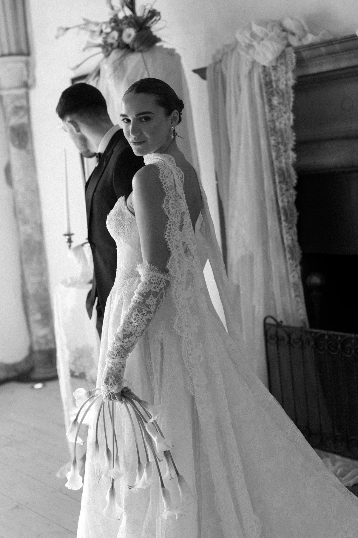 Woman in lace bridal gown looks over her shoulder while walking with a man in a tuxedo. Soft, neutral background. She has a calm, poised expression.