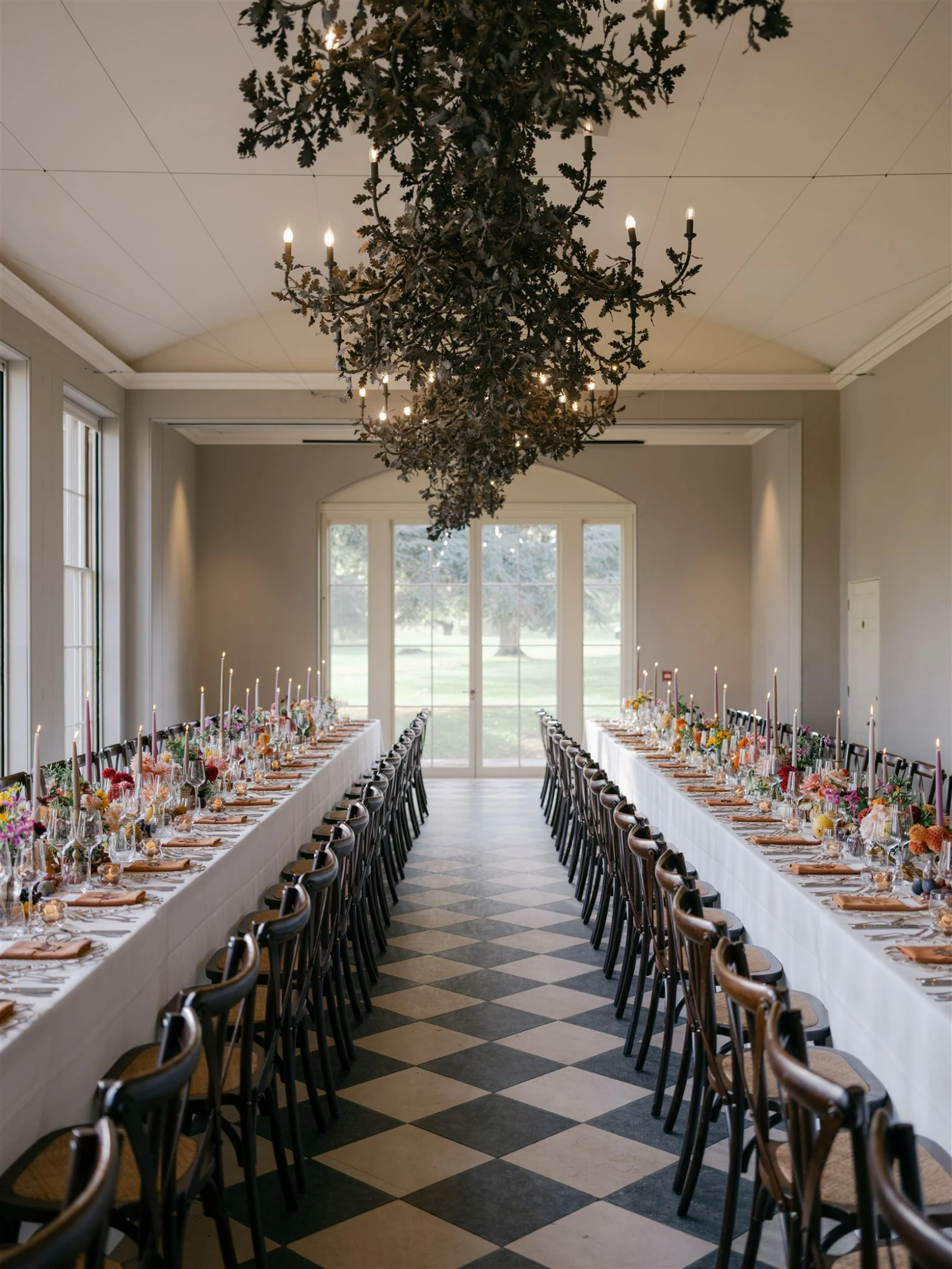 Wedding tables set up in a softly lit room with a black and white checkered floor and a grand black chandelier.