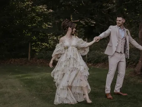 Bride in a flowing white dress and groom in a beige suit hold hands and smile in a wooded garden. Wedding photography in Suffolk by Mikaela Jade.