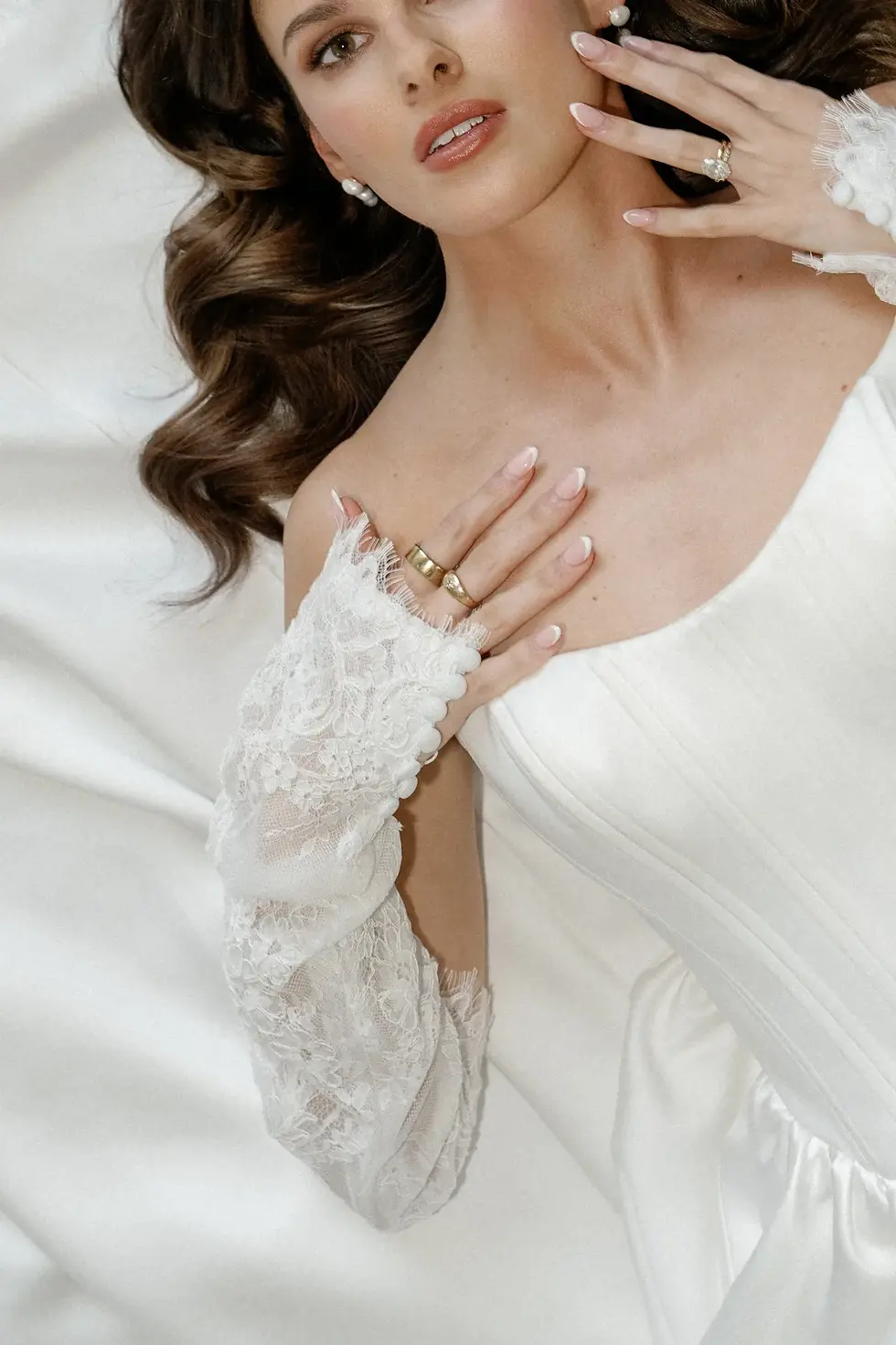 A woman in a white lace dress lies on satin fabric, showcasing rings and pearl earrings; the mood is serene and elegant.