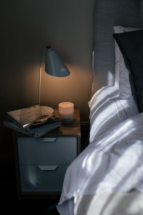 Books, a lamp, and a candle on a bedside table beside white bedding.