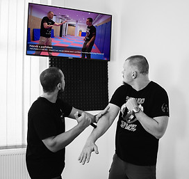 Two men practicing an online self-defense course from a tv on the wall