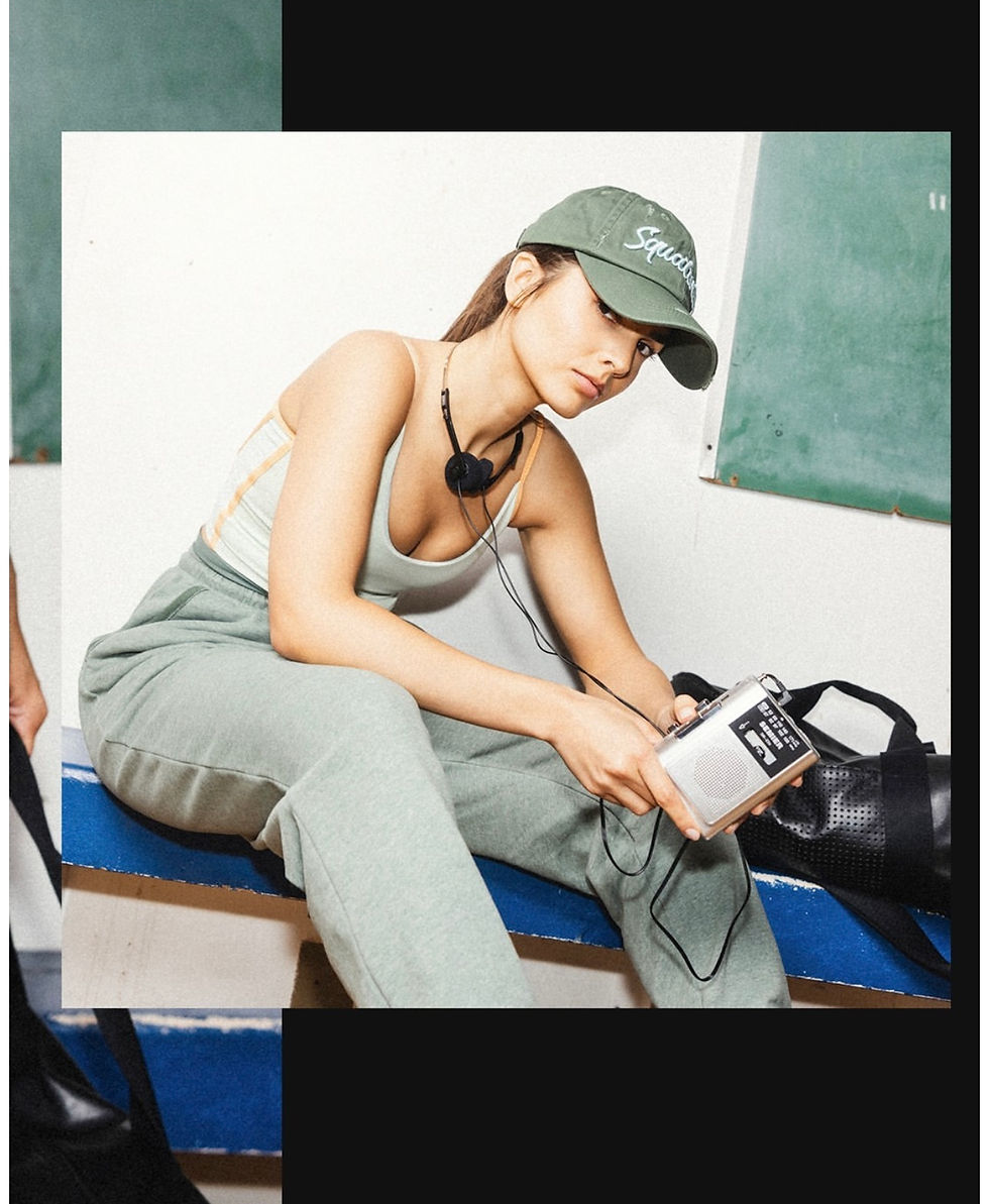 Woman in green cap and outfit listens to a portable radio. She sits on a blue bench, with a chalkboard in the background, calm expression.