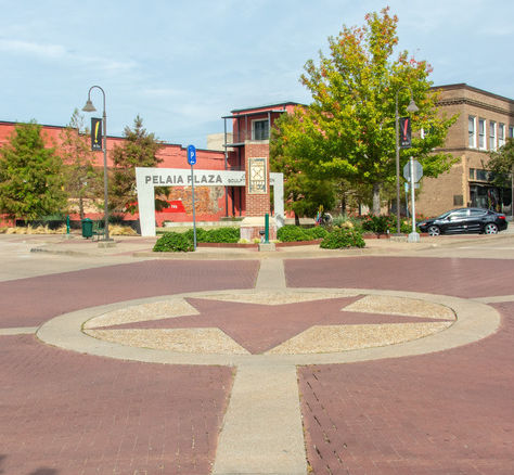 Brick plaza with a star mosaic, storefronts and trees, and a PELAIA PLAZA sign under a blue sky.