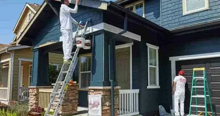 Painters working on the exterior of a blue house using ladders and paint tools.
