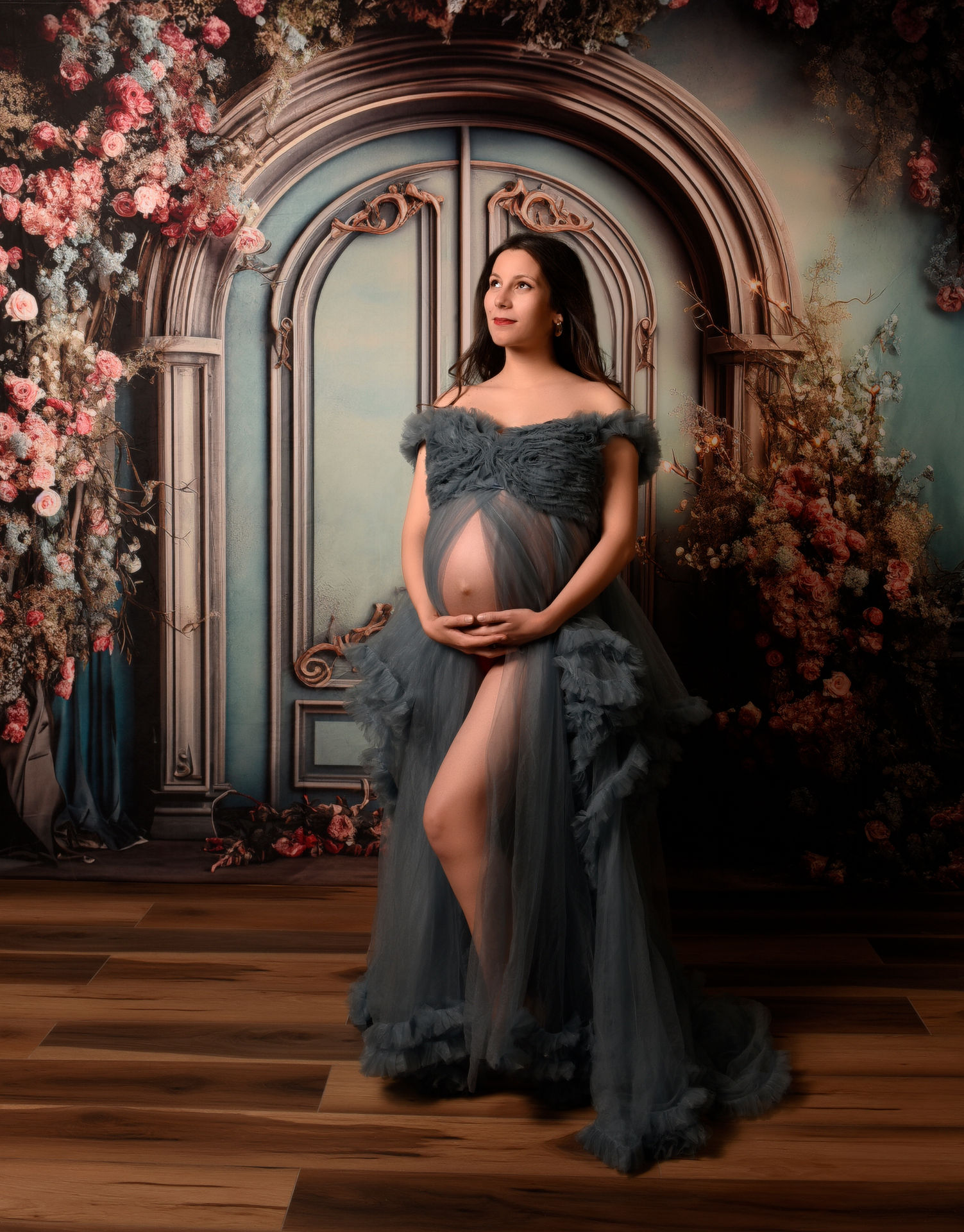 Pregnant woman in ruffled blue gown posing in floral studio.
