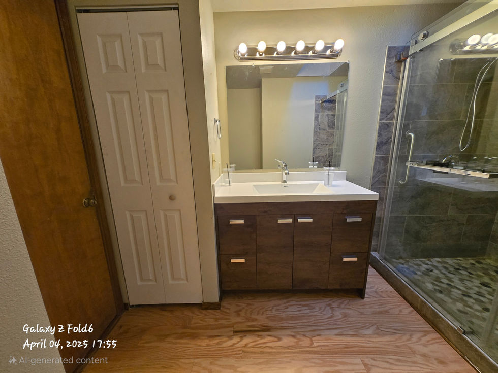 Front view of the bathroom vanity area: a dark wood vanity cabinet with white countertop and sink is centered below a mirror and light fixture, with a white bi-fold closet door to the left and the shower edge to the right.