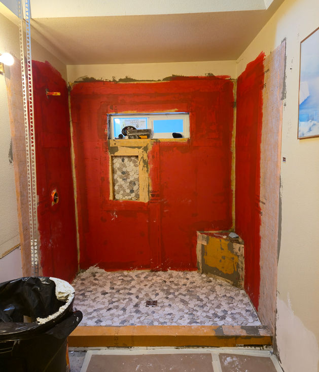 Shower stall walls coated in red RedGard waterproofing membrane, with pebble tile installation underway on the floor and inside a recessed wall niche. A corner seat structure is visible in the lower right.