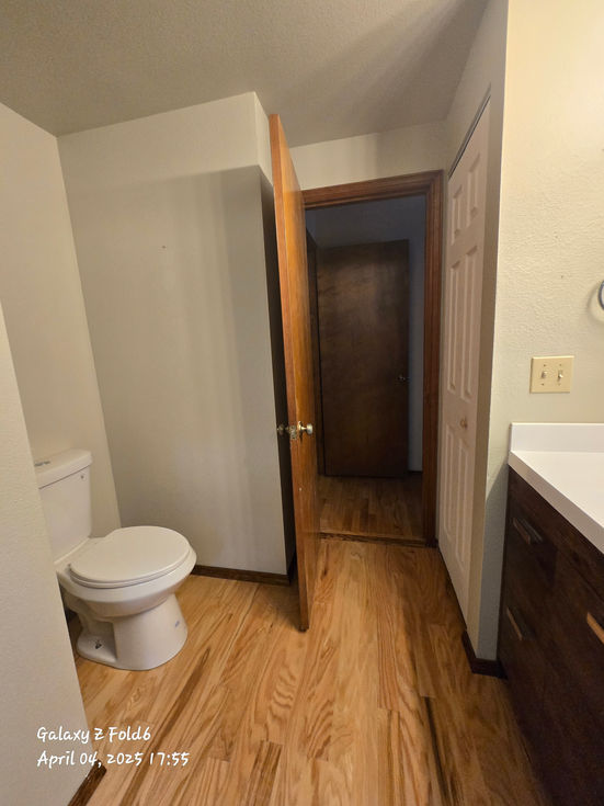 Bathroom view showing the new light wood flooring, a white toilet installed in an alcove on the left, and partial view of a dark wood vanity and white closet door on the right.