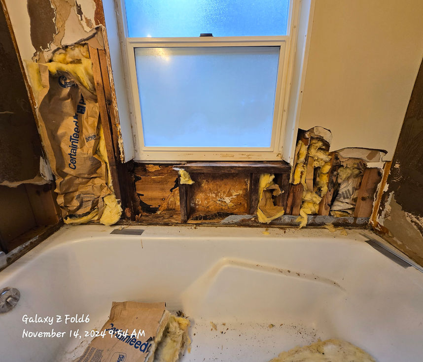 Open wall cavity below a frosted bathroom window showing dark, water-damaged wooden studs and discolored, exposed insulation above a bathtub.