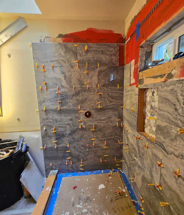 Shower wall being tiled with large grey tiles featuring a round cutout for the shower valve; numerous tile leveling clips are in place. The back wall with niche and window is visible to the right.