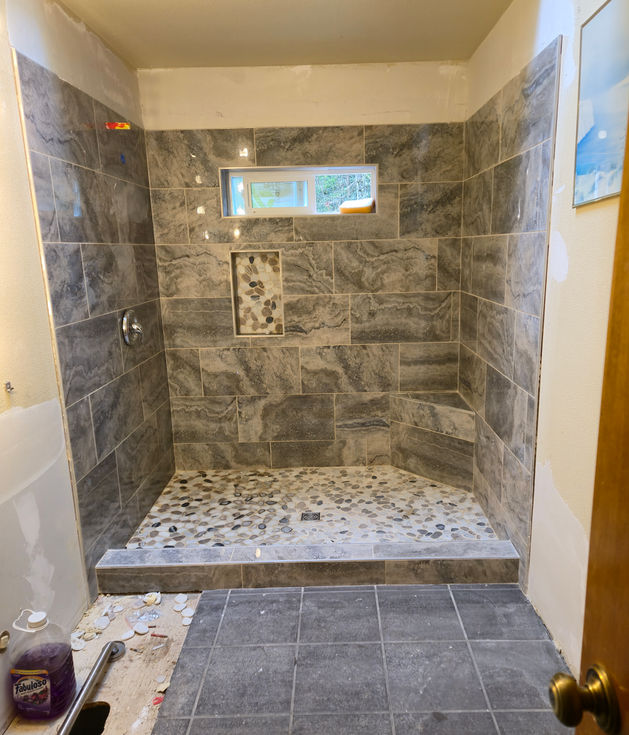 Fully tiled and grouted walk-in shower featuring large grey wall tiles, a light pebble tile floor, tiled niche below the window, and a tiled corner bench. Chrome shower valve trim is visible on the left wall.