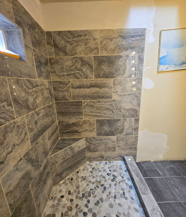 Fully tiled and grouted back wall and corner bench of the walk-in shower, using large grey stone-look tiles. The pebble tile floor is visible in the foreground.