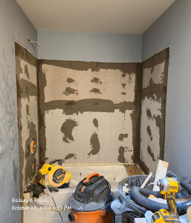 A bathroom shower alcove with cement board walls. Dark gray thin-set mortar is applied over all seams and screw holes, preparing the surface for liquid waterproofing. Tools and equipment are visible in the foreground.