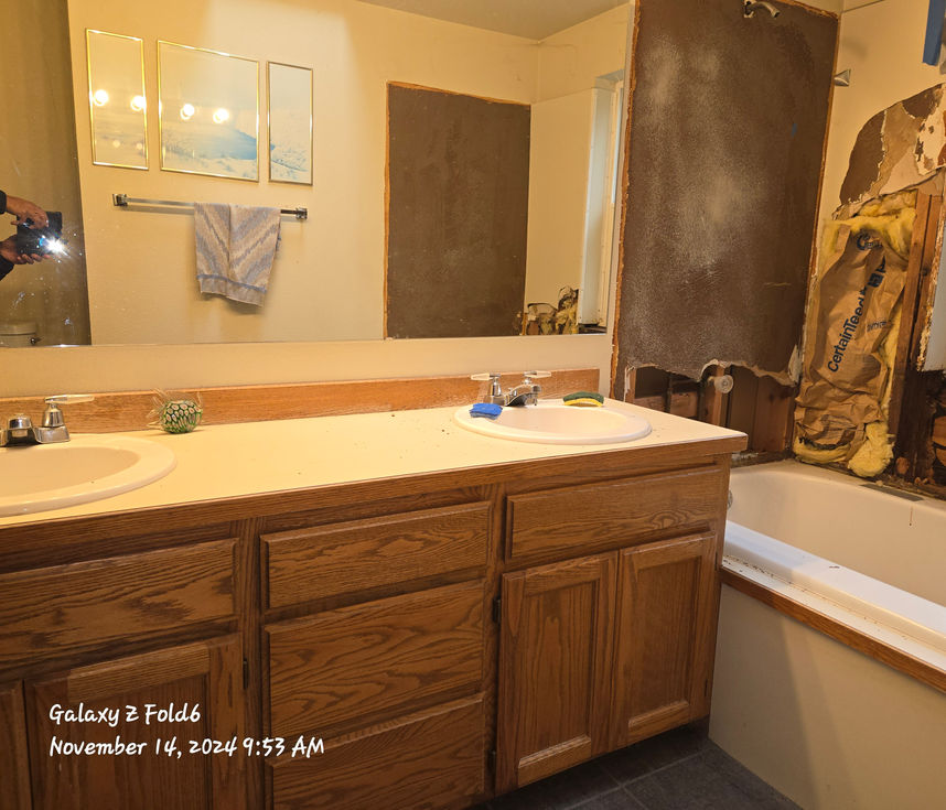 Bathroom prior to renovation featuring a large oak-finish double vanity, laminate countertop, and large mirror, with the damaged wall near the tub visible on the right.