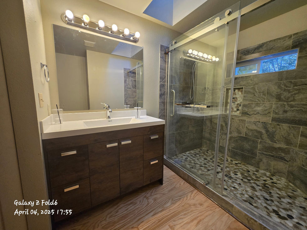 Renovated bathroom showing a dark wood vanity with a white countertop and sink, a large mirror, a contemporary light fixture, the adjacent glass shower enclosure, and light wood flooring.