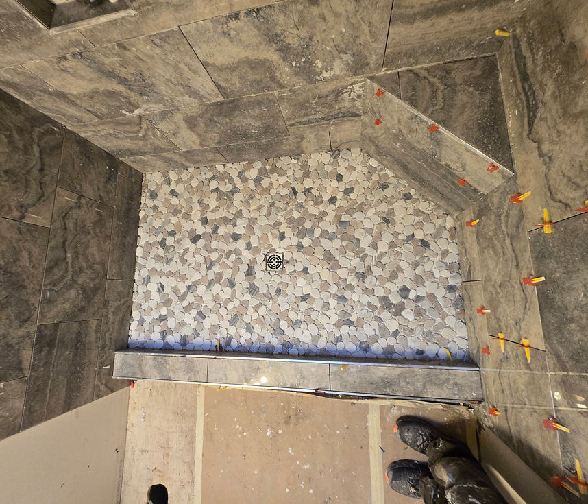 Top-down view of a walk-in shower with completed light-colored pebble tile floor, central square drain cover, tiled curb, and a corner bench tiled with large grey tiles.