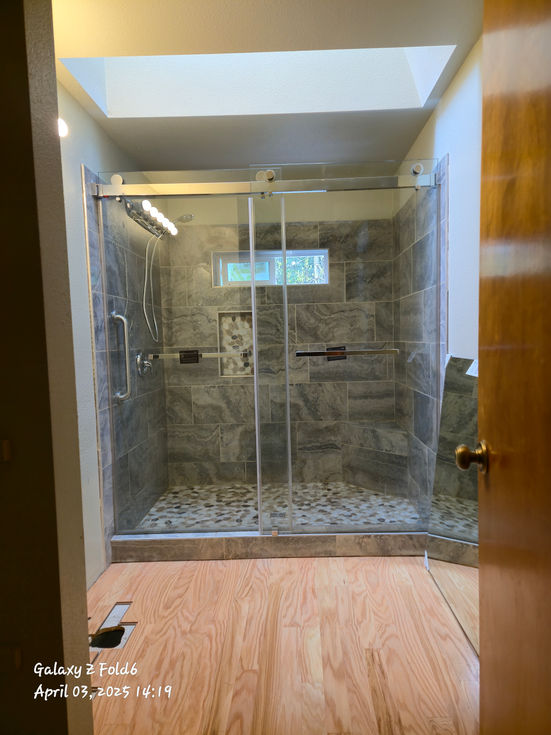Bathroom floor covered in newly installed light-colored engineered wood planks extending from the foreground to the completed shower enclosure in the background.