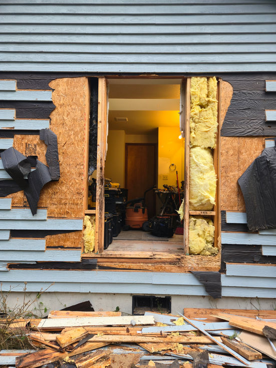 Large rectangular opening in the exterior wall of a house with blue siding, revealing interior wall framing and insulation; demolition debris is on the ground below.