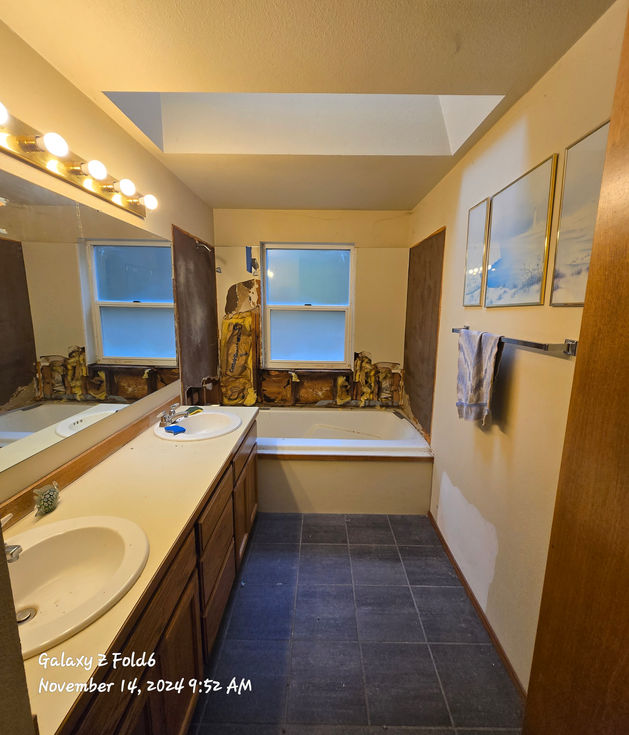 Overview of the bathroom before renovation: double vanity on left, tub at the far end under a window with wall damage visible below it, dark floor tiles, and a skylight above.
