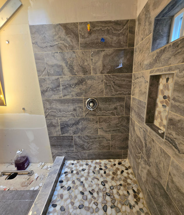 The left wall of the shower, fully tiled with large grey stone-look tiles and grouted, featuring the installed chrome shower valve trim. Pebble tile floor visible below.