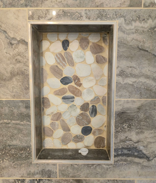Close view of a recessed shower niche with a pebble tile mosaic on the back surface and framed with large grey tiles on the sides and sill.