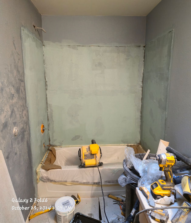 A bathroom shower alcove with walls coated in a light green, smooth liquid waterproofing membrane. The new white tub is visible below, covered for protection. Tools are in the foreground.