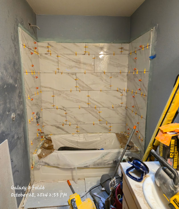 A bathroom shower alcove with newly installed white rectangular tiles with gray veining on the walls. Numerous yellow and red tile leveling spacers are visible between the tiles, ensuring even grout lines. The white tub below is covered for protection.