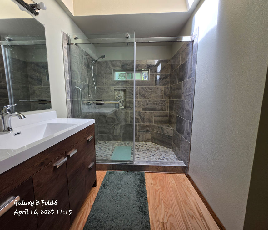 Fully renovated bathroom showing vanity on the left, glass shower on the right, light wood flooring with a dark bath mat in front of the shower, and a lighter mat inside the shower.