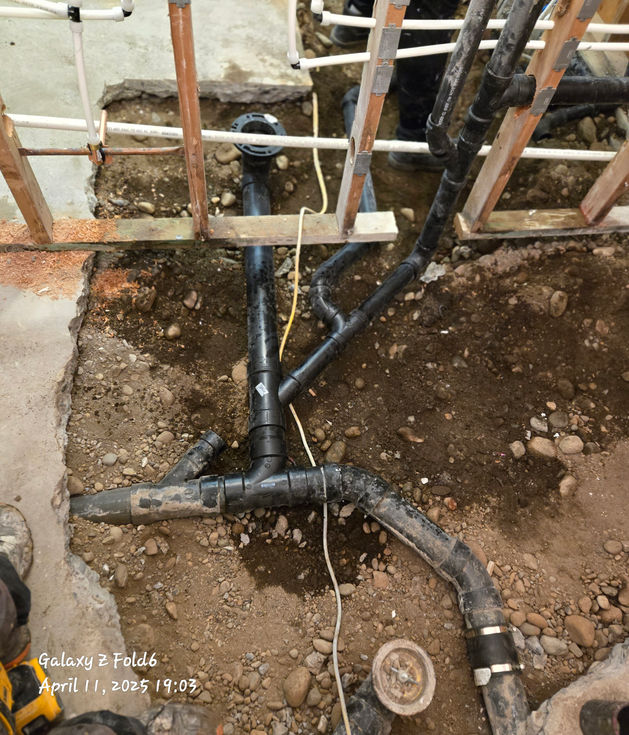 View of a newly installed black PVC/ABS underground plumbing waste line network laid in excavated dirt under a concrete slab.