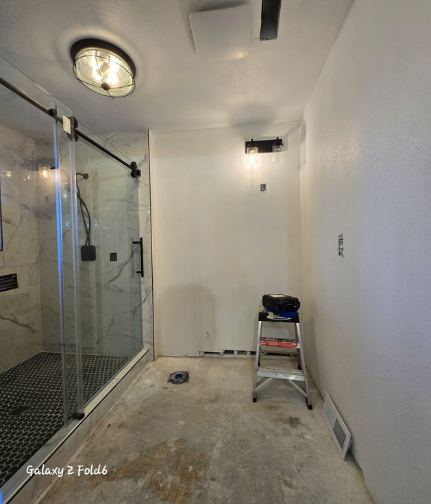 Modern master bathroom with a glass-enclosed walk-in shower with black hardware, newly painted walls, black light fixtures, and an unfinished concrete floor.