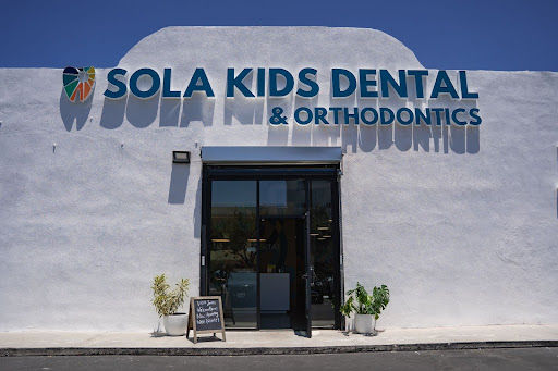 White storefront of Sola Kids Dental & Orthodontics with glass entrance, potted plants, and blue sky.