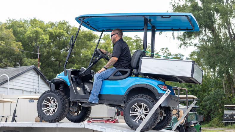 A team member guiding a bright blue custom lifted golf cart up the loading ramp of a transport trailer.