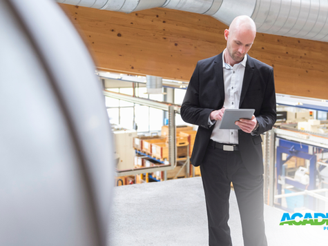 Man in suit using tablet in industrial setting with wooden ceiling and windows. "ACADEMY PUMP & MOTOR" logo at bottom right.