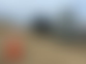 Construction site with large machinery and orange cones in a dirt area. Overcast sky and autumn trees in the background.