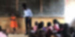 A teacher and a smiling girl in orange stand by a chalkboard in a classroom. Students sit at desks, focusing on the lesson.