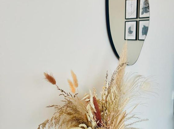 Vaas met gedroogde planten op een oppervlak tegen een witte muur, met een onregelmatig gevormde spiegel erboven en vier ingelijste afbeeldingen in een vierkant patroon aan de rechterkant.